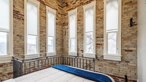 Un dormitorio con pared de ladrillo, ventanas grandes, una cama y una manta azul.