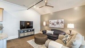 Una sala moderna con una televisión de pantalla plana, un ventilador de techo, un sofá, un reposapiés y un cuadro en la pared.