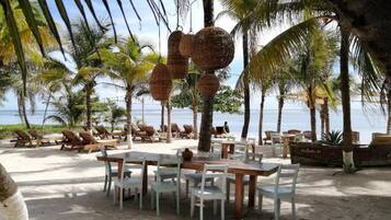 A beachside restaurant with outdoor seating, palm trees, and a view of the ocean.