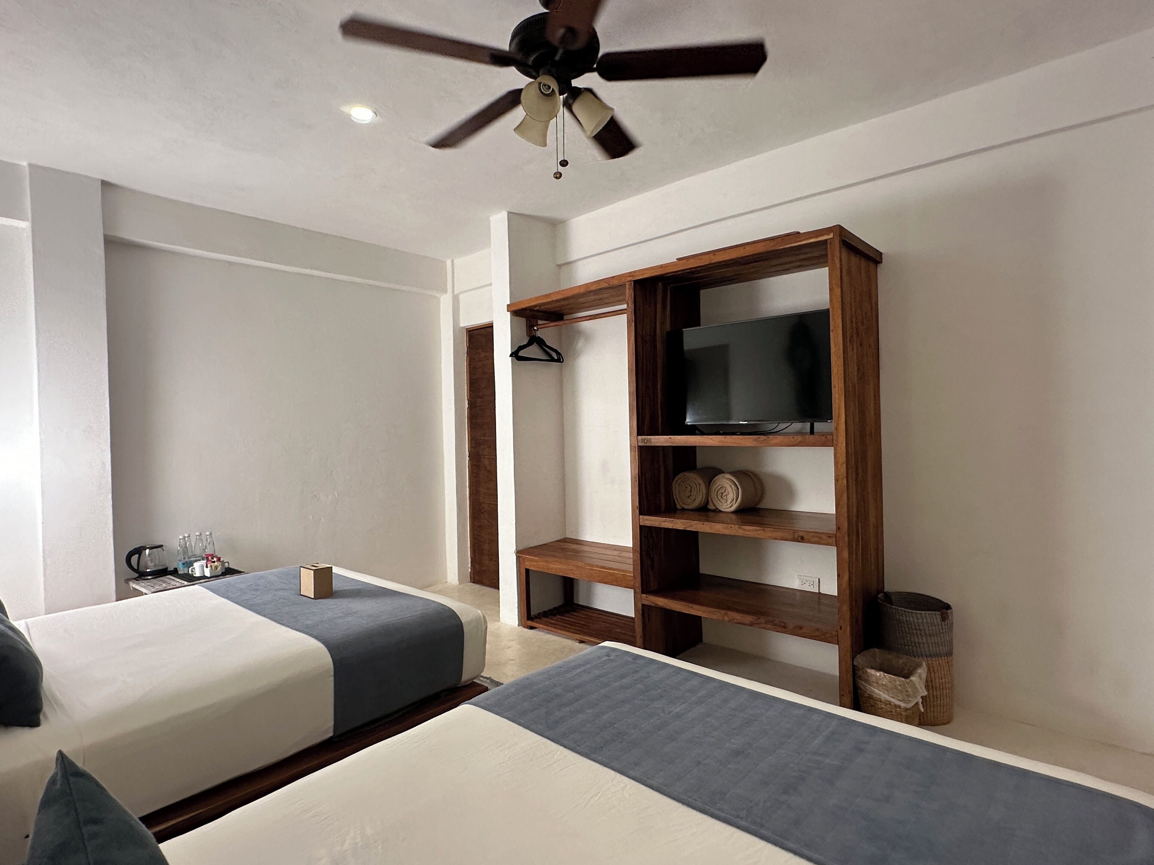 A hotel room with two beds, a ceiling fan, a TV on a wooden shelf, and a small table with a kettle and cups.