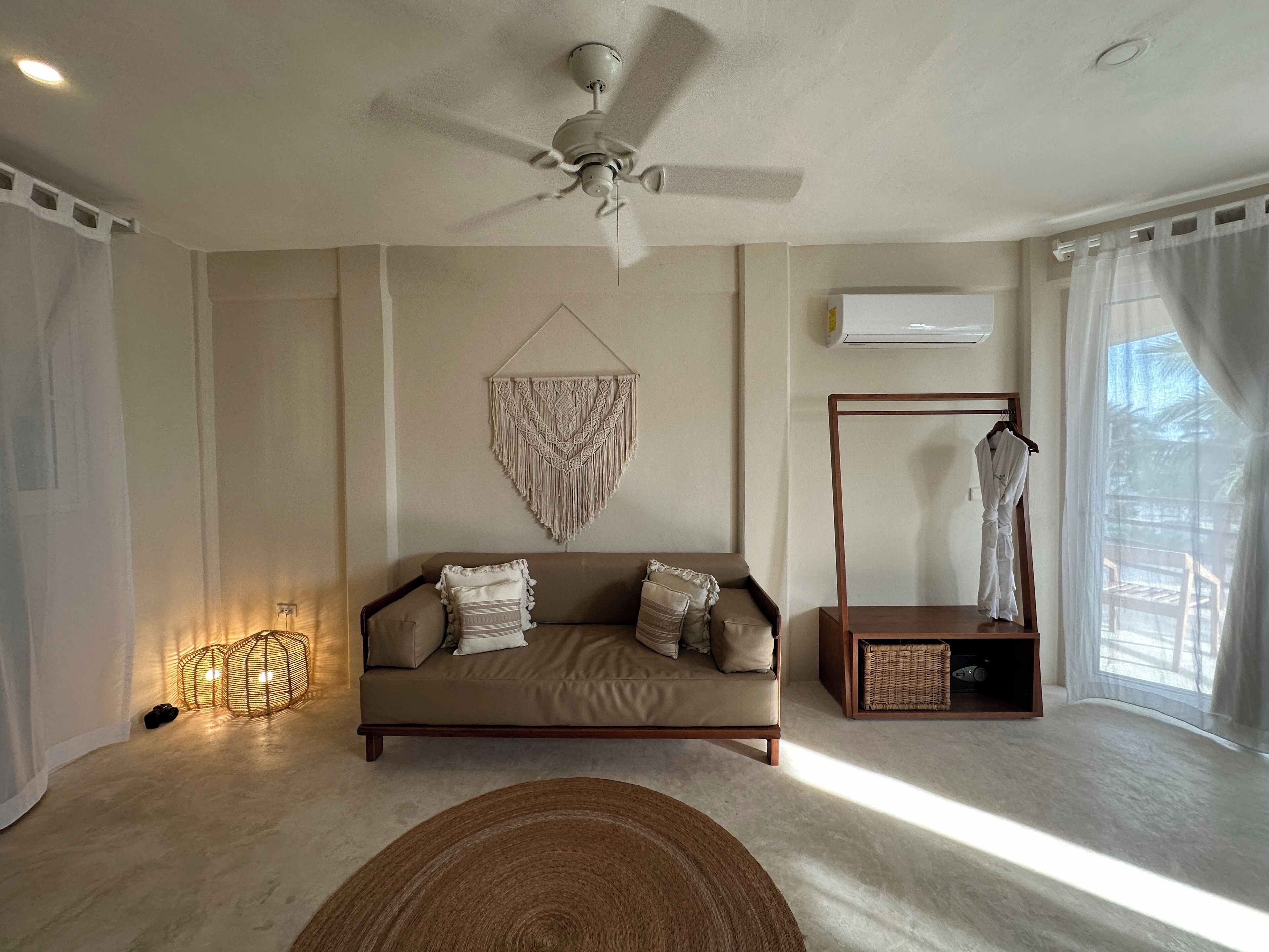 A modern living room with a beige sofa, a ceiling fan, a macrame wall hanging, a wooden side table, and a large window with sheer curtains.