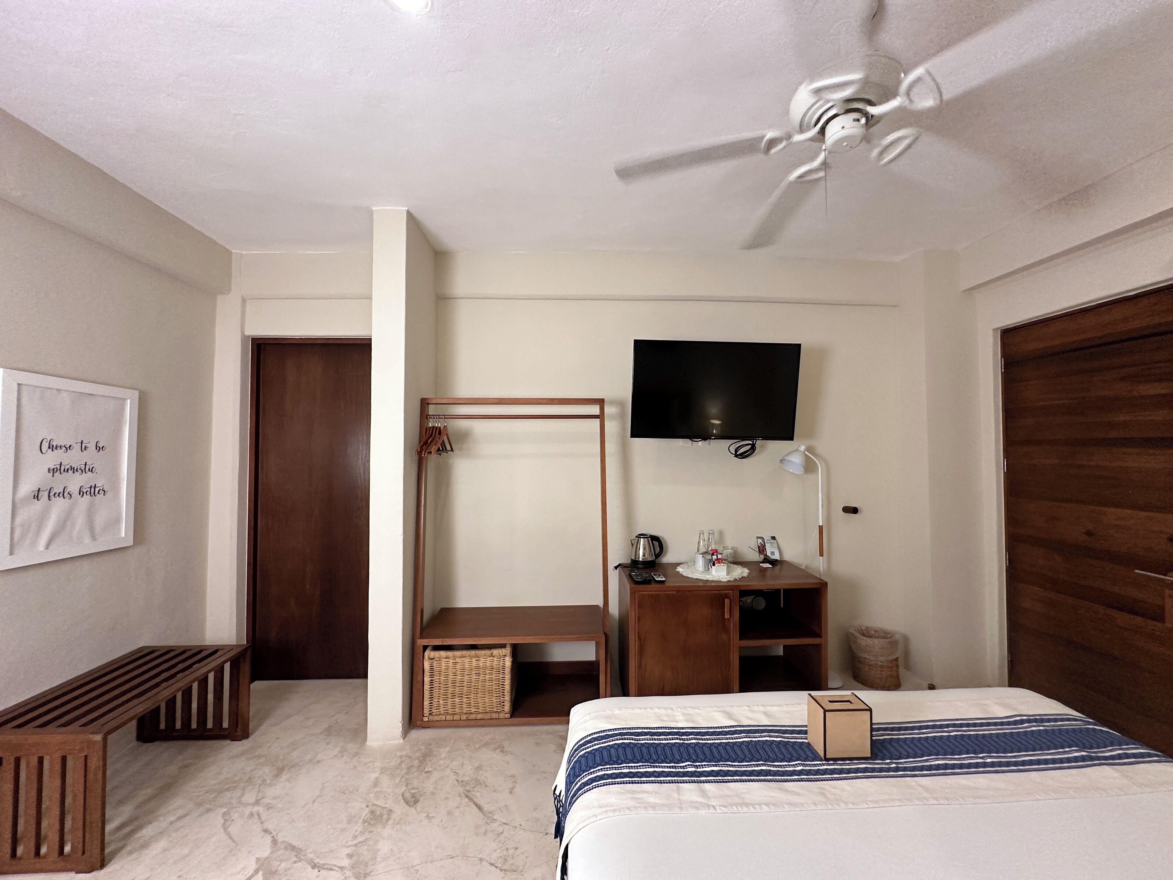 Quarto com cama, ventoinha no teto, televisão, escrivaninha com abajur e guarda-roupa de madeira.