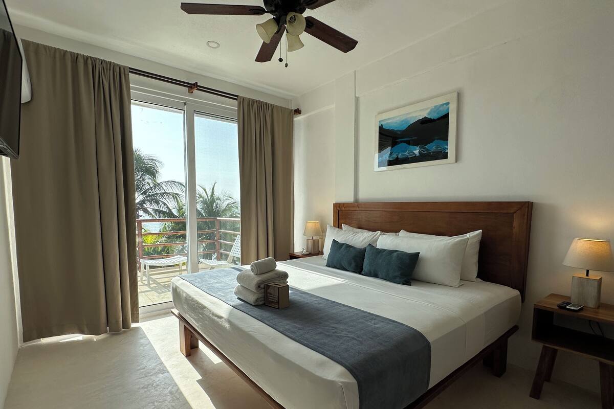 Ein Hotelzimmer mit einem großen Bett, einem Deckenventilator, einem Gemälde und Blick auf Palmen durch das Fenster.