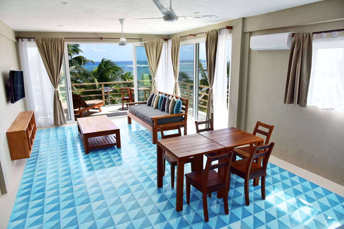 Ein Zimmer mit blauem geometrischem Boden, einem Holzesstisch mit Stühlen, einer Couch mit Kissen, einem an der Wand montierten Fernseher und einem Balkon mit Blick auf das Meer und Palmen.