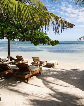 Una playa con sillones de madera bajo una palmera, agua azul cristalina y una orilla arenosa.