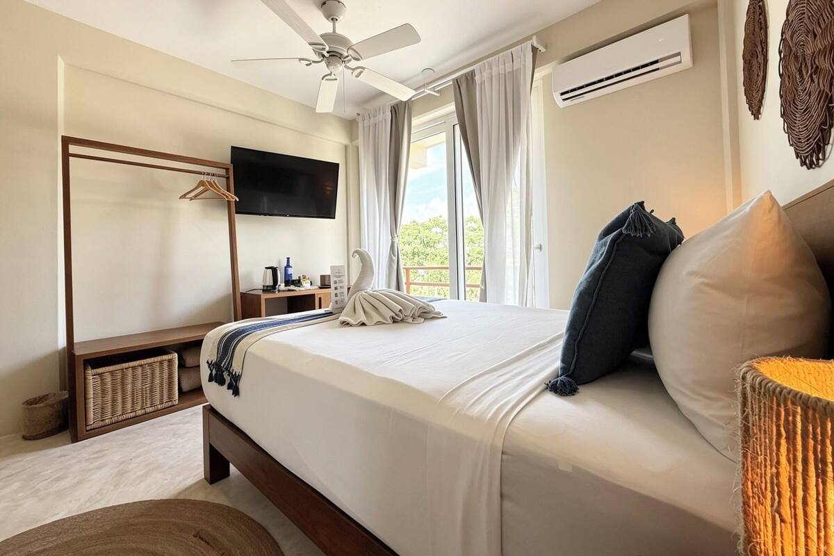 Ein Schlafzimmer mit Bett, Deckenventilator, Fernseher, einem Fenster mit Vorhängen und Blick auf einen Balkon.
