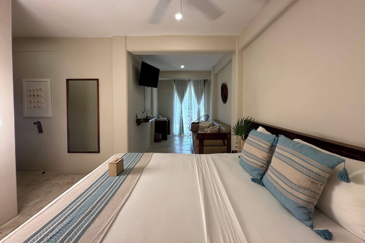 Ein Hotelzimmer mit einem großen Bett, einem Deckenventilator, einem an der Wand montierten Fernseher und Blick auf den Flur.