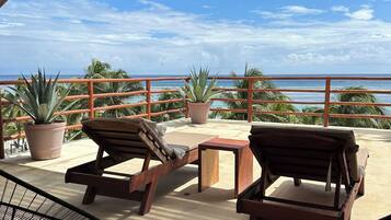 A covered outdoor seating area with lounge chairs and a table, overlooking a beach with palm trees and the ocean.