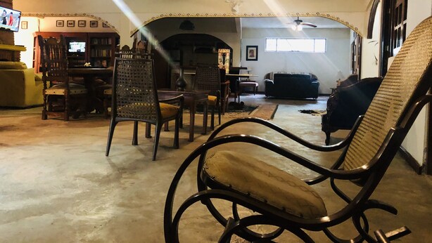 Un salon spacieux avec un plafond haut, des ventilateurs de plafond et une cheminée. On y trouve plusieurs fauteuils et un canapé, ainsi qu’un téléviseur fixé au mur.