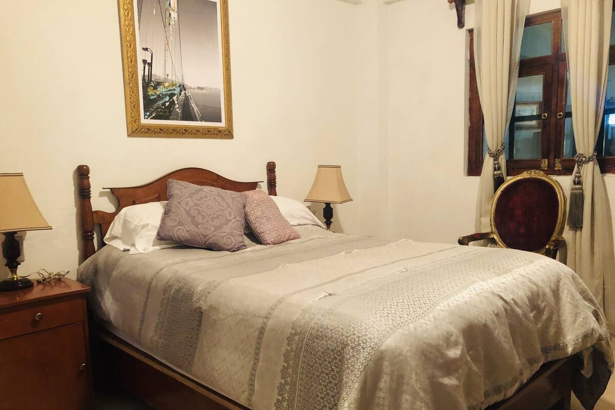A neatly made bed with a patterned bedspread and two pillows, a wooden nightstand with a lamp, and a mirror on the wall.