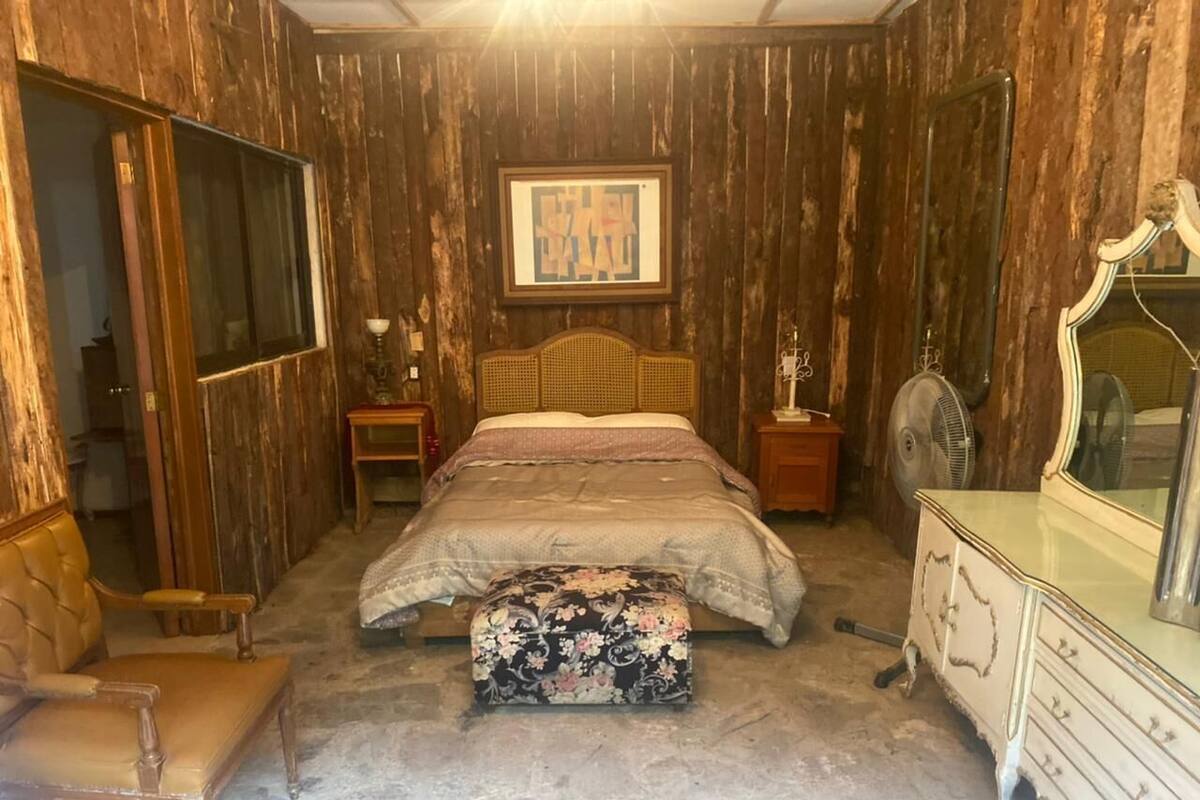 A bedroom with a wooden paneled wall, a bed with a floral bedspread, a wooden chair, a dresser, and a mirror.