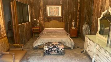 Un dormitorio con pared de paneles de madera, una cama con cubrecama floral, una silla de madera, un aparador y un espejo.