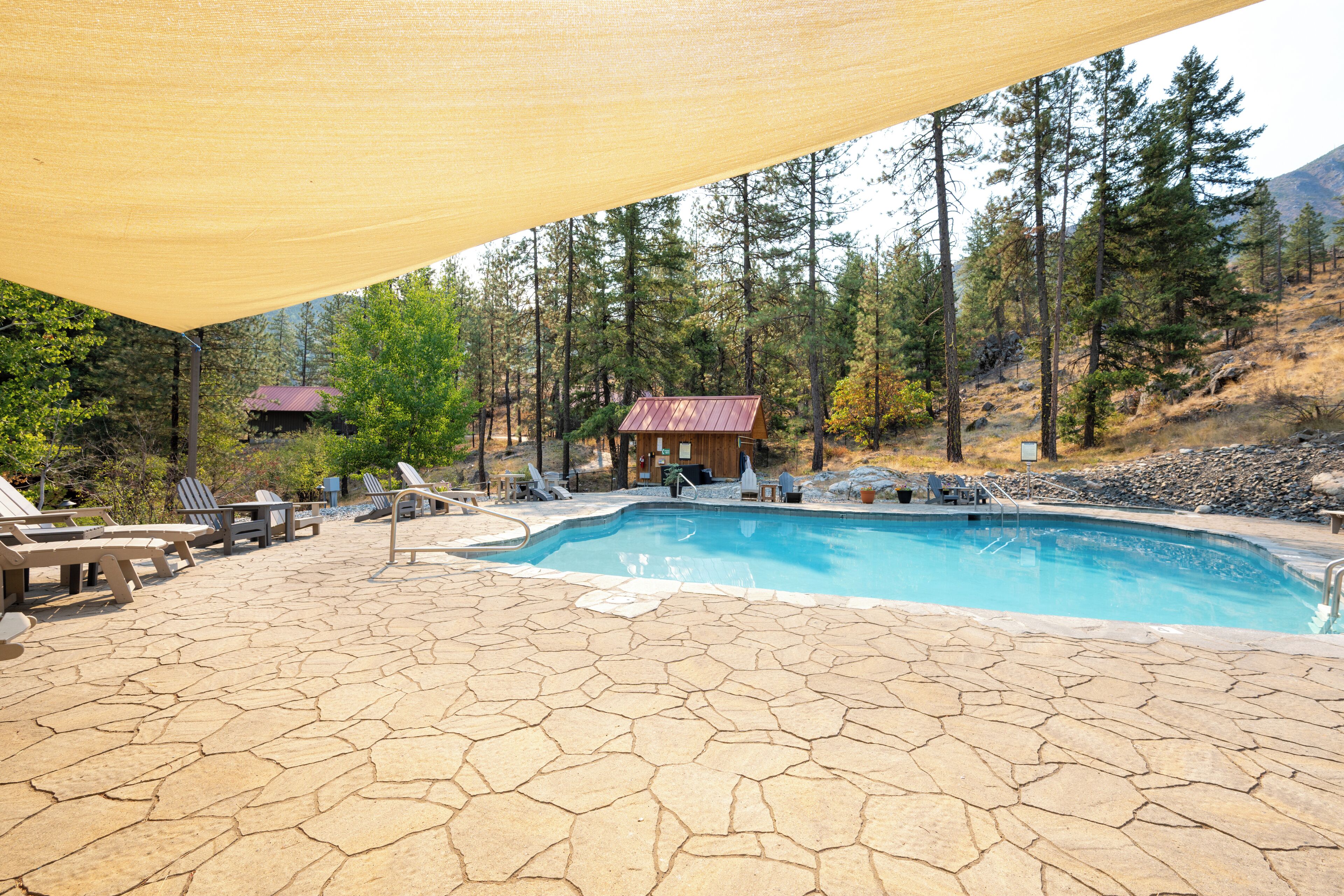 A swimming pool with lounge chairs and a covered area, surrounded by trees and a mountain in the background.