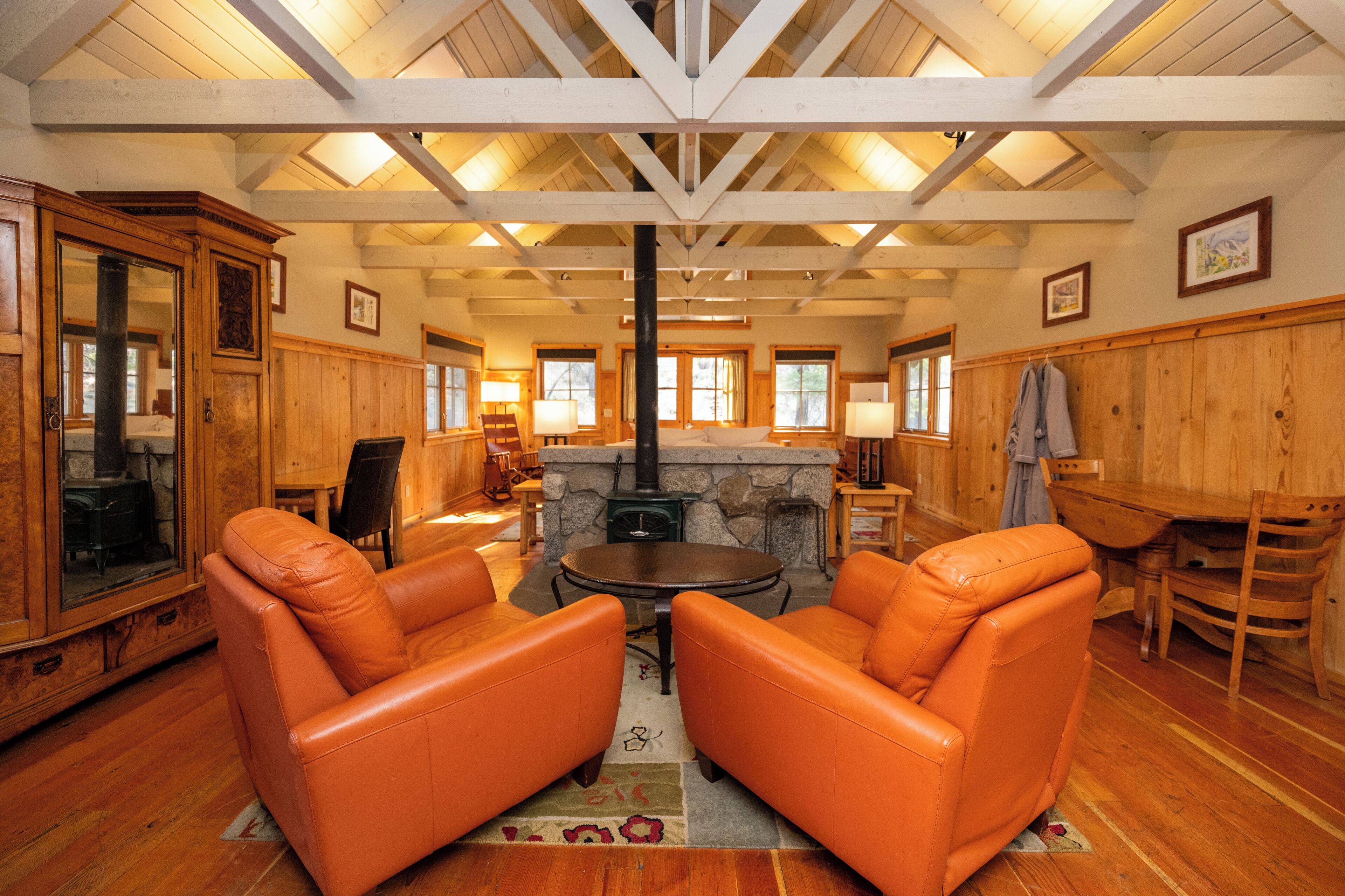 Un salon chaleureux aux murs en bois et à la cheminée en pierre, agrémenté de fauteuils en cuir orange et d’une table en bois.