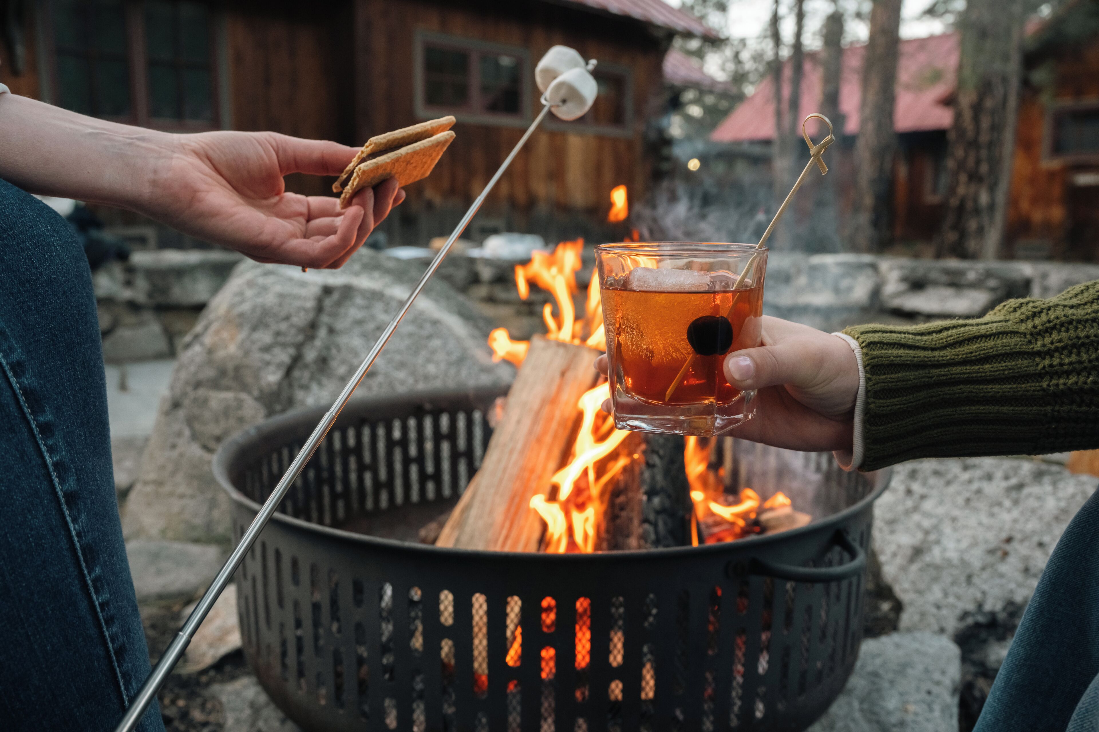 A person holding a marshmallow over a campfire, another person holding a glass with a drink, and a basket of wood.