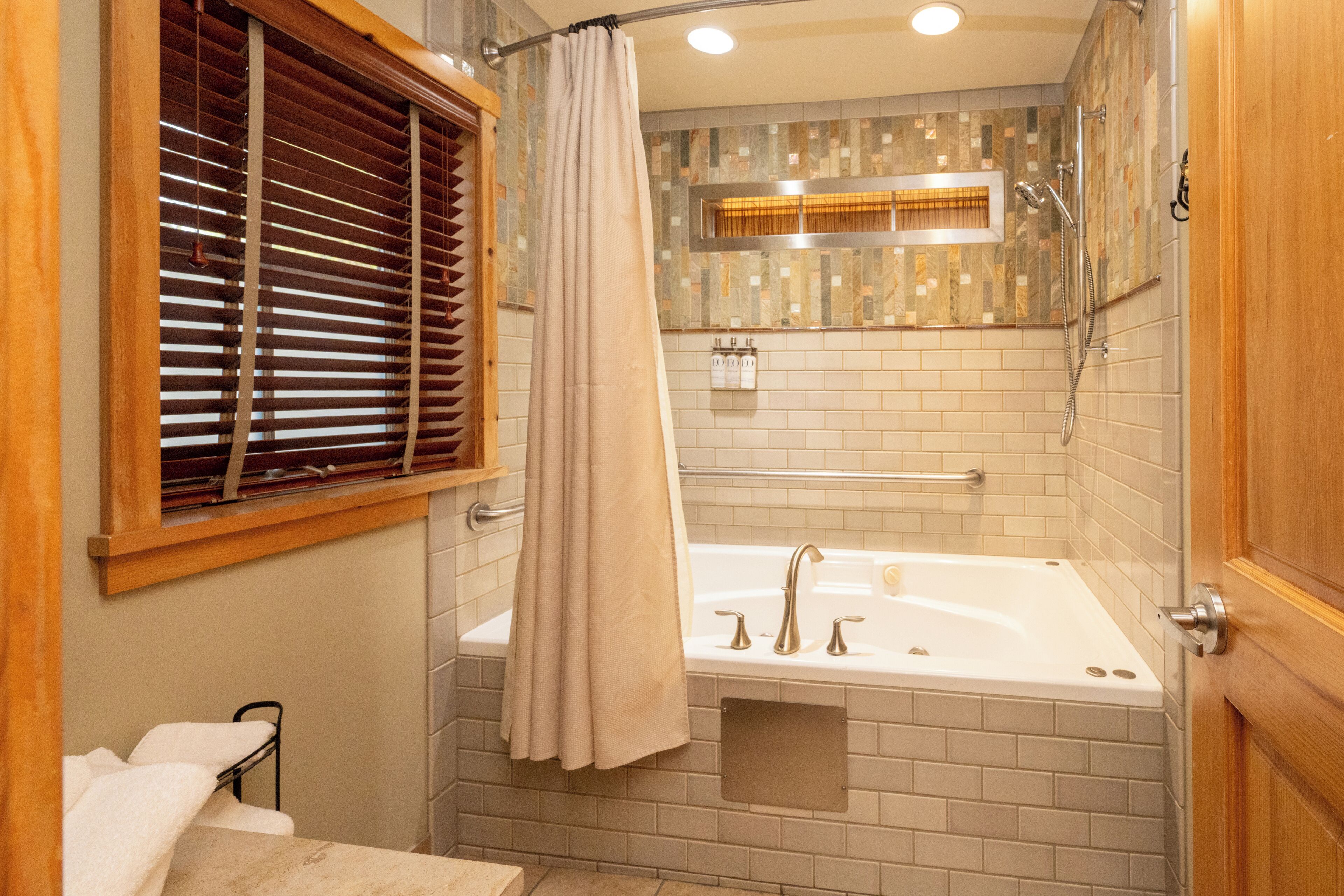 A modern bathroom with a bathtub, shower, and window with blinds.