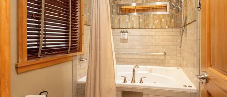A modern bathroom with a bathtub, shower, and window with blinds.