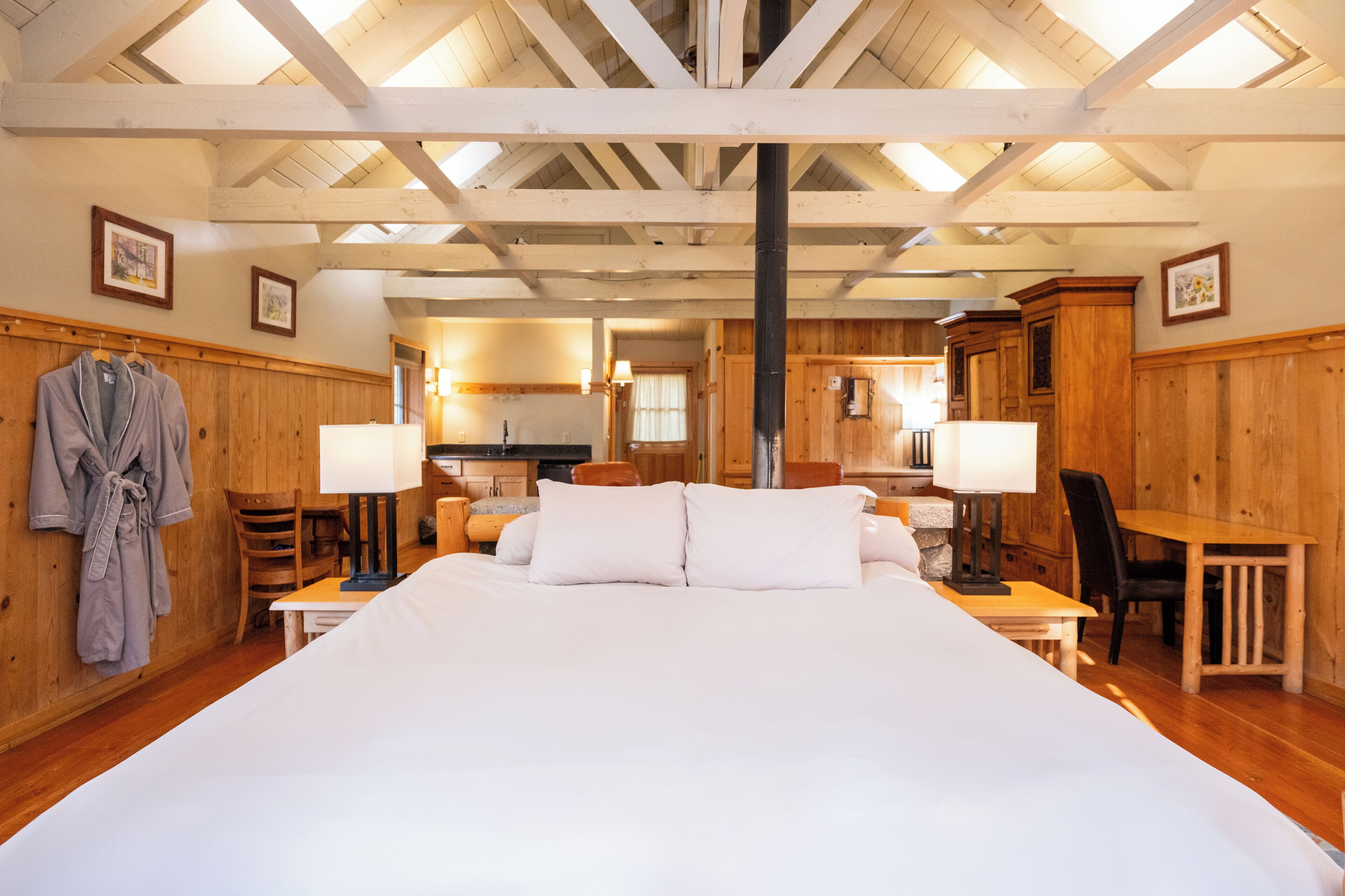 A large bed with white linens in a wooden room with a high ceiling and exposed beams. There are two chairs and a table with lamps.