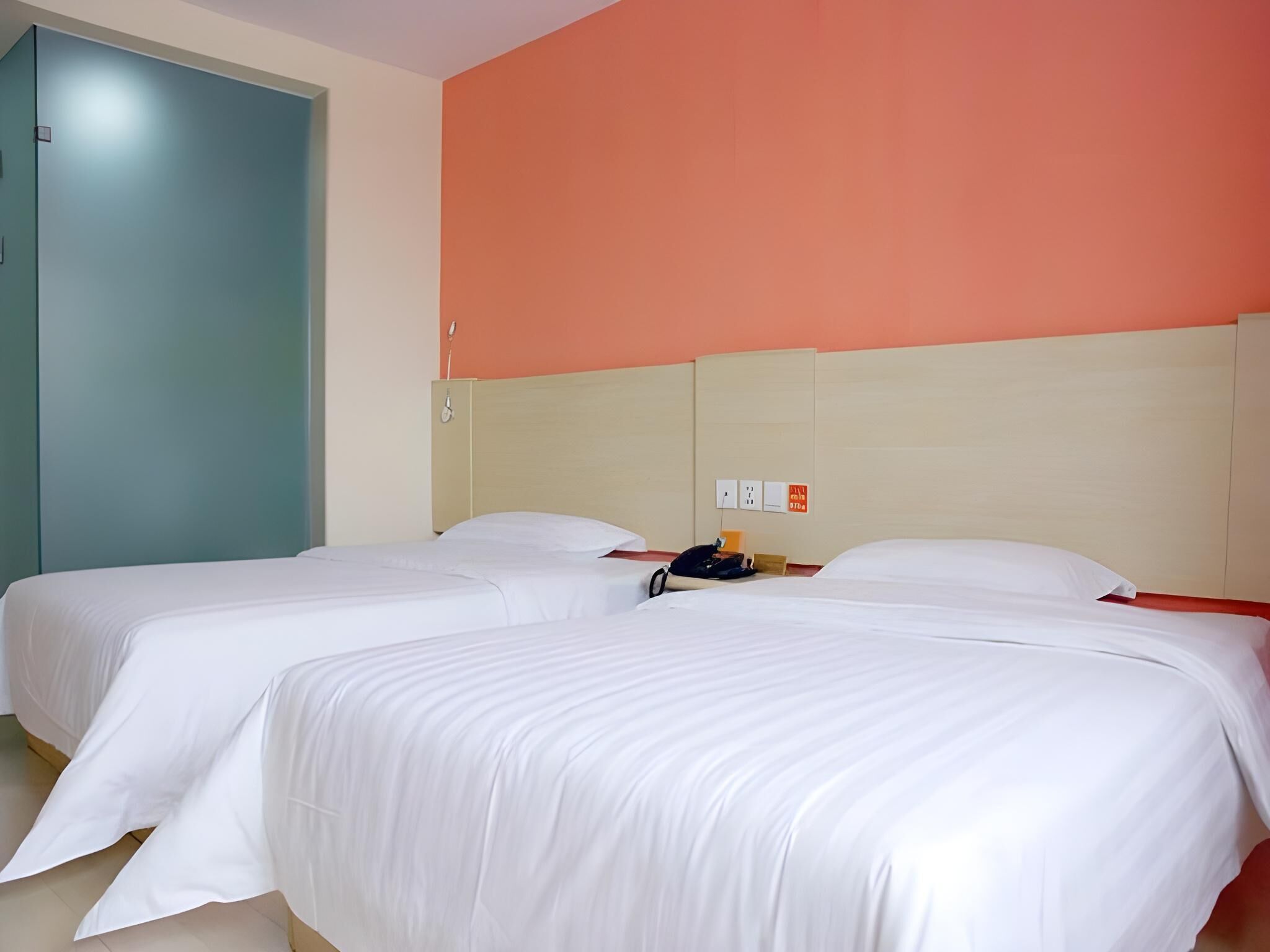 A hotel room with two single beds, a wooden headboard, a teal door, and a wall with a power outlet and a phone.