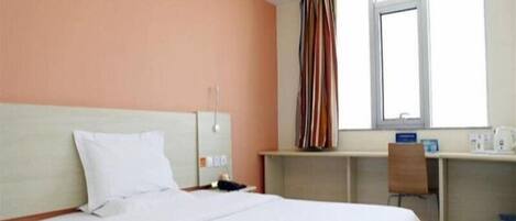 A hotel room with a bed, a desk, a chair, a window with curtains, and a small table with a kettle.