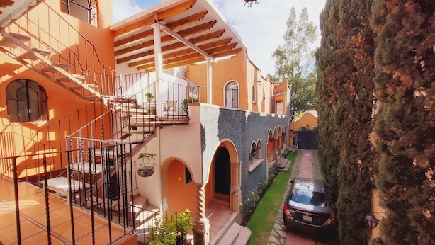 A courtyard with a staircase leading to a balcony, a car parked nearby, and a building with arched windows and a tiled roof. - Villa Alfonsina (Mexico City)