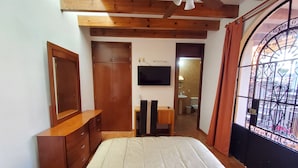 A bedroom with a bed, wooden wardrobe, TV, desk, and a bathroom visible through an open door. - Villa Alfonsina (Mexico City)