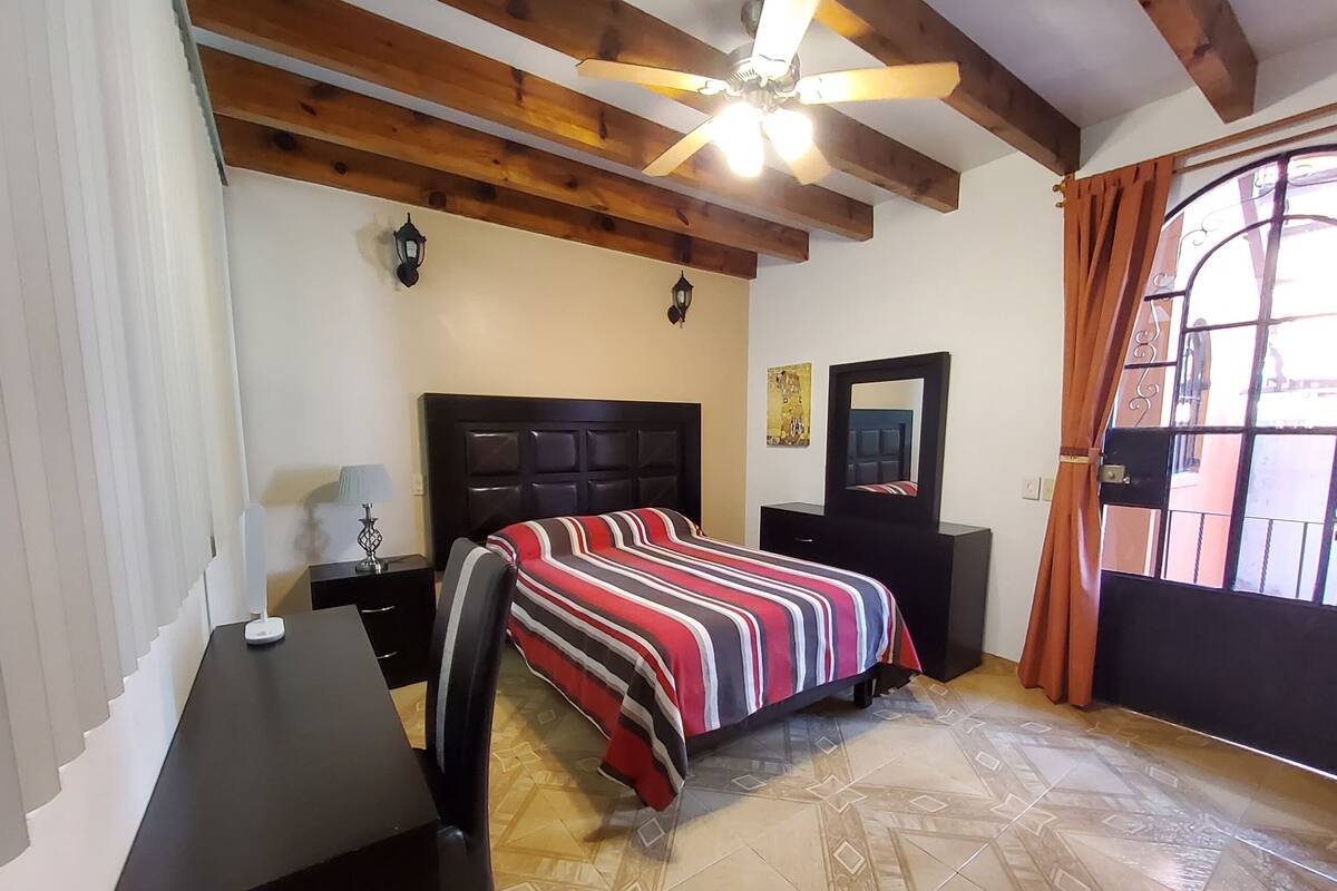 Un dormitorio con cama, escritorio, silla, ventilador de techo, mesita de noche, cómoda, espejo y ventana con cortinas.