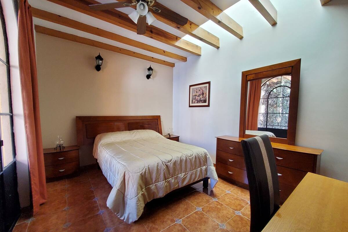 Un dormitorio con techo de madera, una cama, un escritorio, una silla y una ventana con marco decorativo.