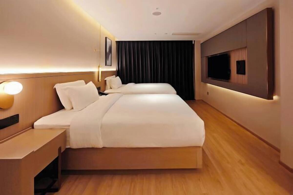 Ein Hotelzimmer mit zwei Betten, einem an der Wand befestigten Flachbildfernseher, einem Nachttisch und einem großen Fenster mit Vorhängen.