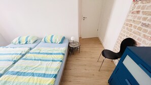 A room with two beds, a small table with a lamp, a chair, and a blue dresser.