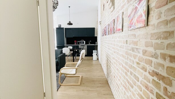 A narrow hallway with a brick wall on one side and a door on the other. There are two white chairs and a small table in the distance.