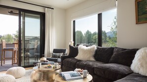 Un salón moderno con un sofá gris oscuro, una puerta de cristal que da a un balcón y vistas a árboles y montañas.