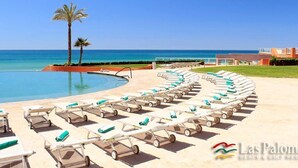 Un espace aménagé au bord de la piscine, avec des chaises longues et un palmier, offrant une vue sur une mer d’un bleu limpide.