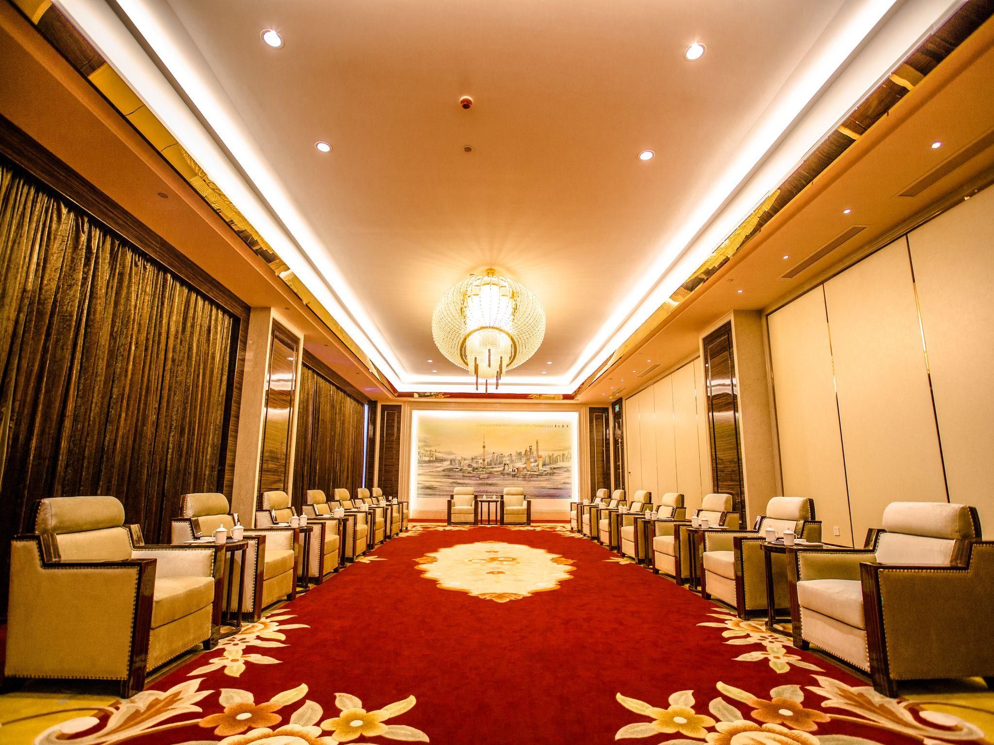 Uma sala de conferências de hotel com tapete vermelho, fileiras de cadeiras, um lustre e um mural da paisagem urbana.