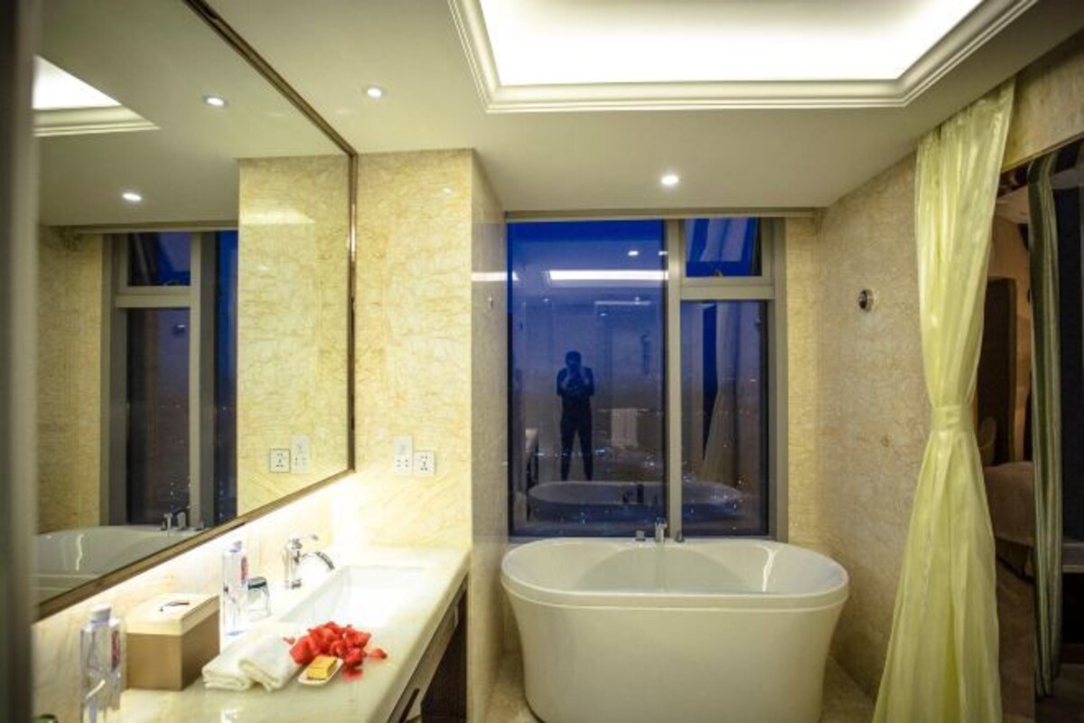 Une salle de bain moderne avec une grande baignoire, un meuble-lavabo avec un miroir et une fenêtre donnant sur l’extérieur, où l’on peut apercevoir une personne.