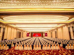 A conference room with rows of chairs, a stage with a red backdrop, and a patterned carpet. - Fuyue Hotel (Shanghai)