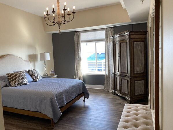 A bedroom with a large bed, a chandelier, a wardrobe, and a window with curtains. - Beautiful Updated Condo In Grand Haven (Grand Haven)