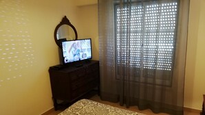 A room with a television on a dark wooden dresser, a patterned rug, and a window with sheer curtains. - Lisbon Coast (Costa da Caparica)