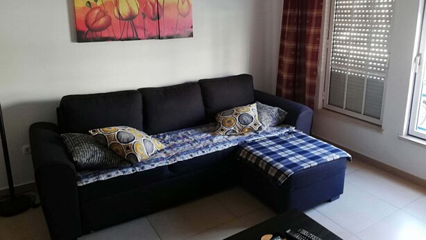A living room with a black sofa, patterned cushions, a blue and white checkered blanket, a painting of flowers, and a window with blinds. - Lisbon Coast (Costa da Caparica)