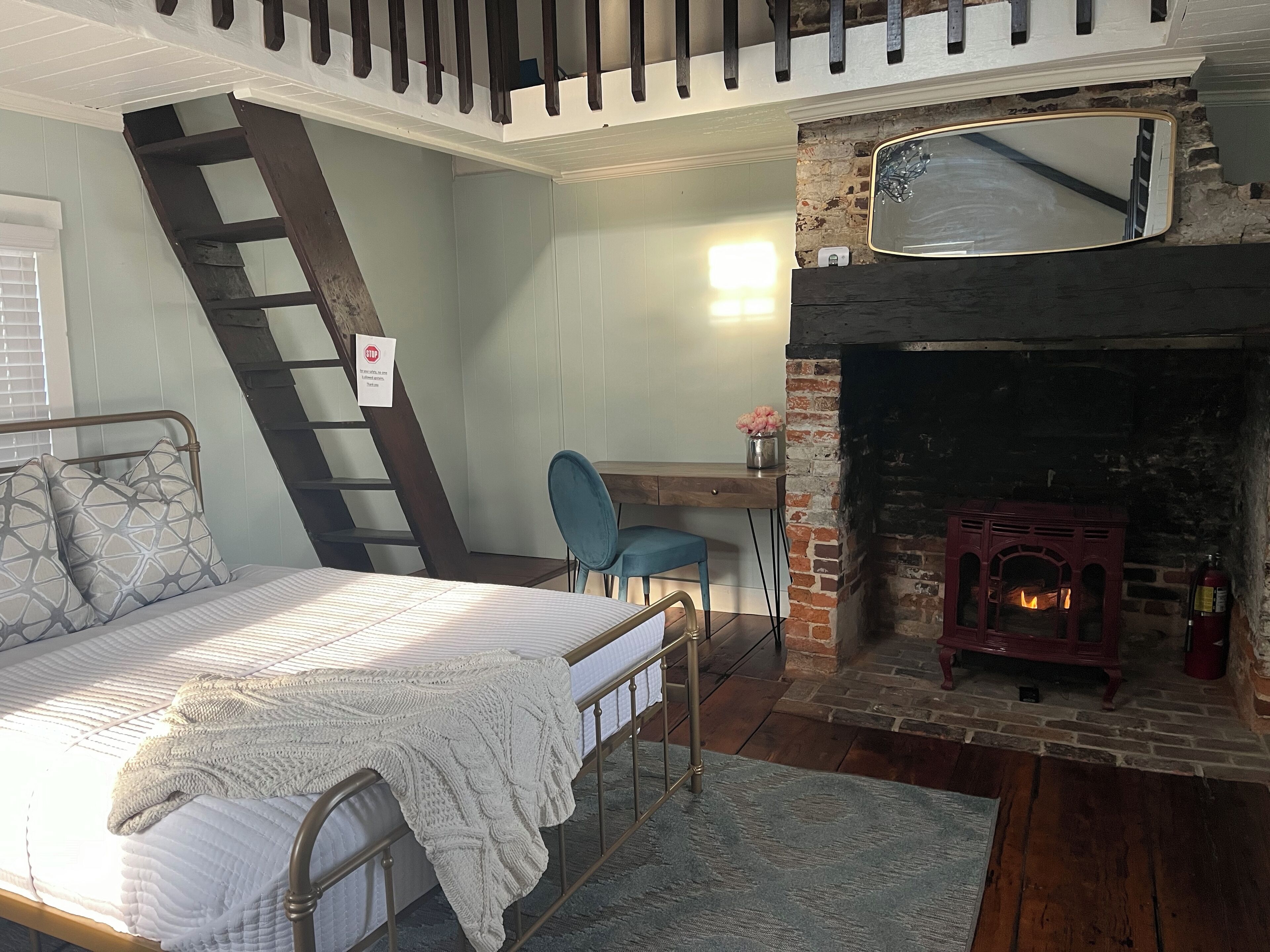 A bedroom with a bed, a desk, a chair, a fireplace, and a staircase.