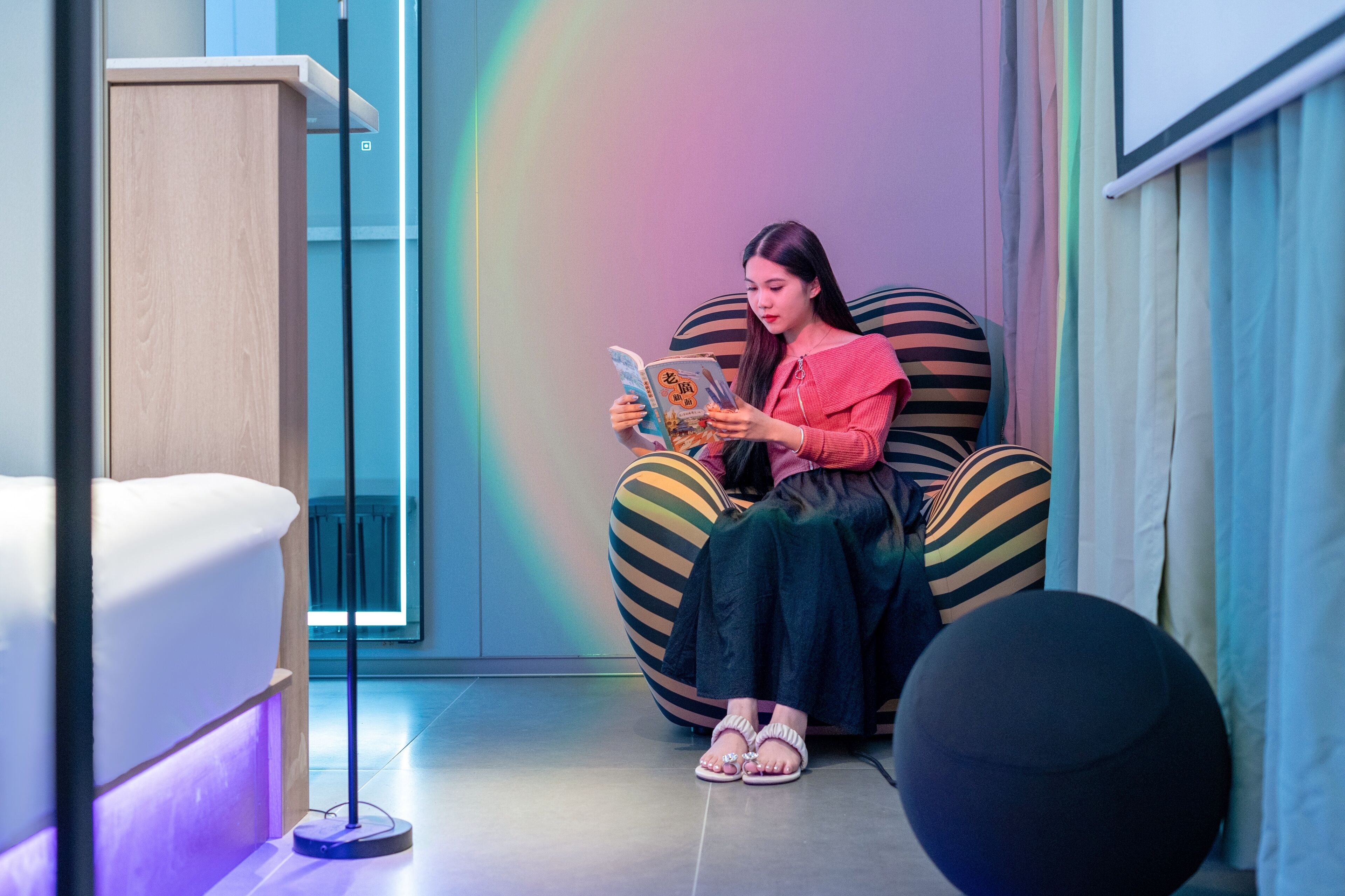 A person sitting on a striped bean bag chair reading a book in a room with a modern design, including a wooden cabinet and a floor lamp.