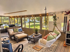 A sunroom with a sofa, chairs, a hanging swing, and a view of a green landscape. -   Peaceful Waterfront Retreat on Three Mile Lake • Bring Your Boat (Port Barre)