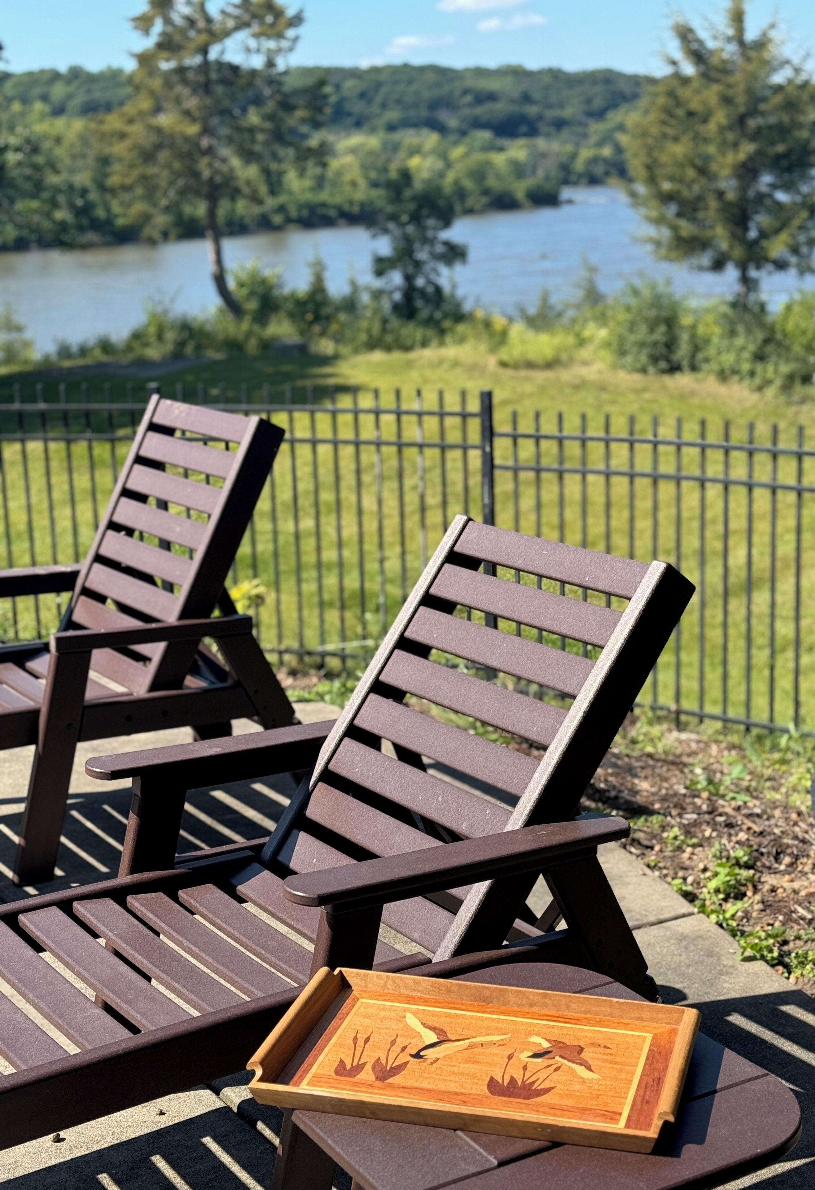 Deux fauteuils de jardin en bois, accompagnés d’une petite table, sont placés sur une terrasse dotée d’une clôture en métal, avec une vue sur le lac en arrière-plan.
