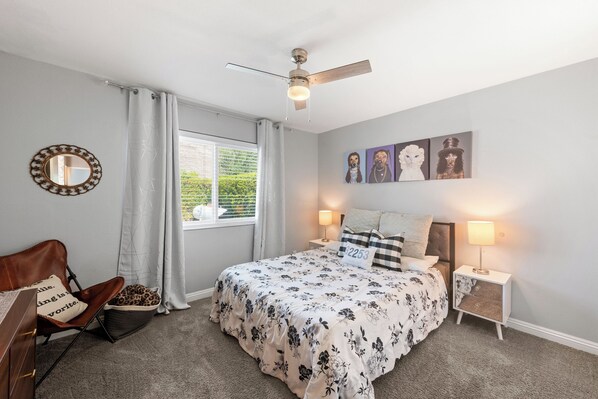 A bedroom with a bed, a chair, a nightstand, a lamp, a ceiling fan, and artwork on the wall.