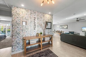 Un interior moderno con papel tapiz floral, un letrero de bienvenida, una consola de madera, un sofá gris y un área de comedor con televisor.