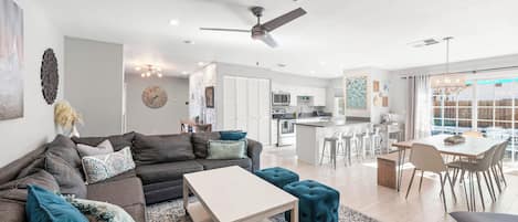 A modern living room with a sofa, ottoman, and coffee table. There is a kitchen area with a dining table and chairs.