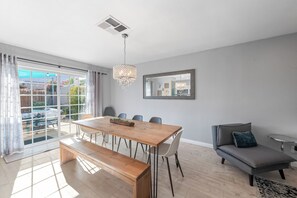 A modern living room with a wooden dining table, a bench, a sofa, and a large window with curtains.