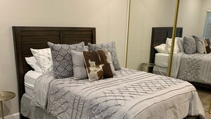 Ein ordentlich bezogenes Bett mit einem gemusterten Überwurf und mehreren Kissen, ein runder Nachttisch und ein großer Spiegel an der Wand.