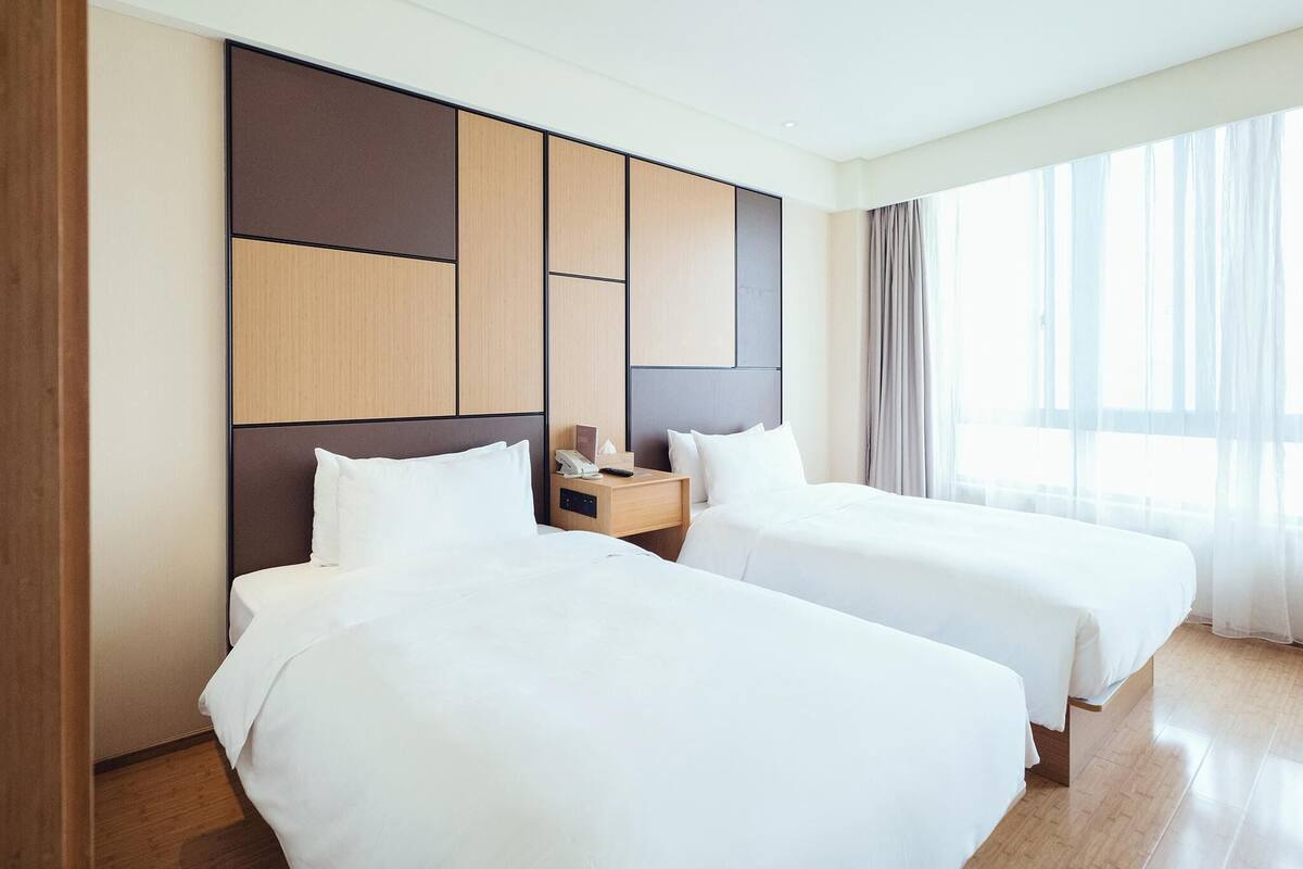 Une chambre d’hôtel avec deux lits, une tête de lit en bois, une table de chevet avec un téléphone et des mouchoirs, et une grande fenêtre avec des rideaux.