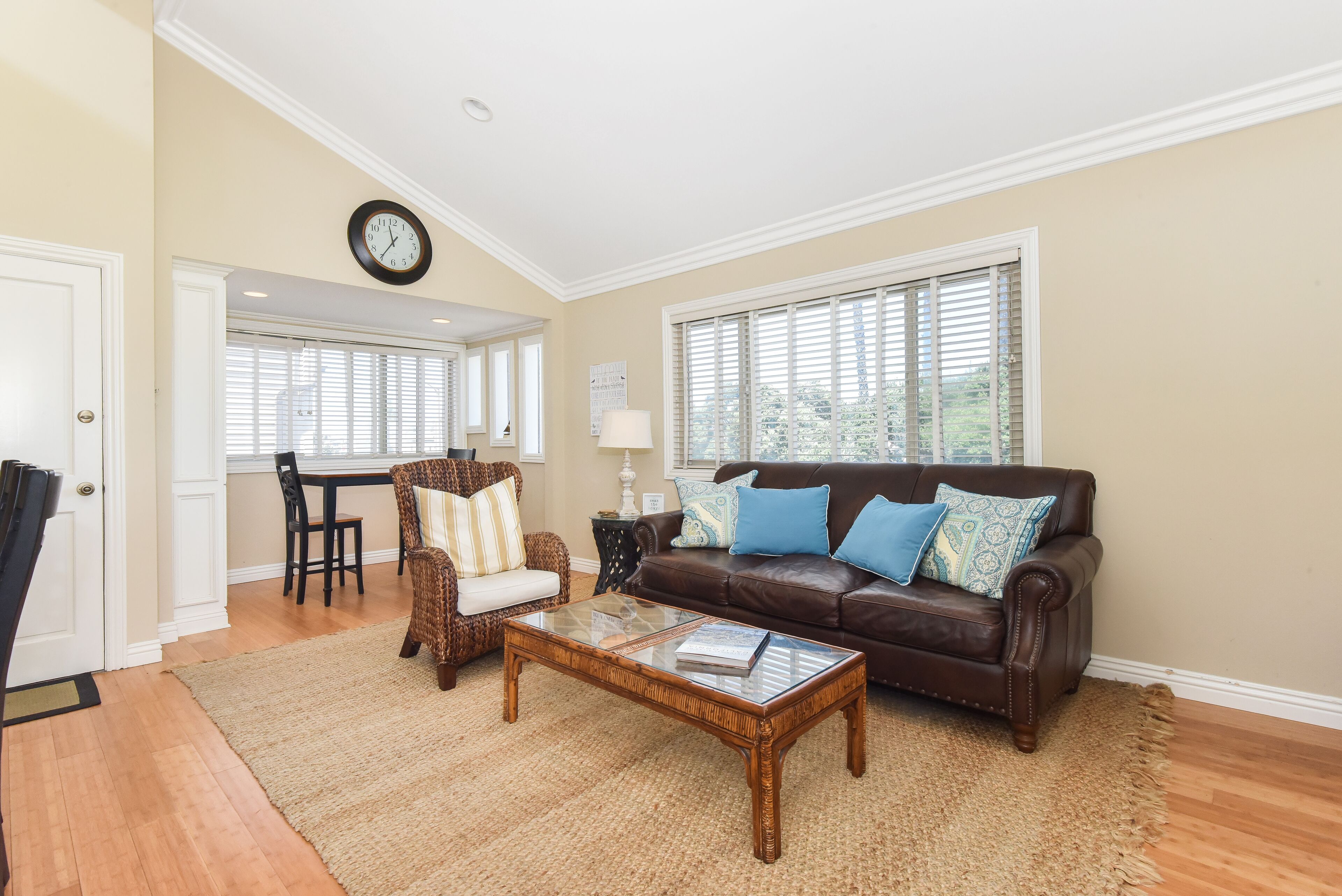 A living room with a brown leather sofa, a glass coffee table, a wicker chair, and a dining table with chairs.